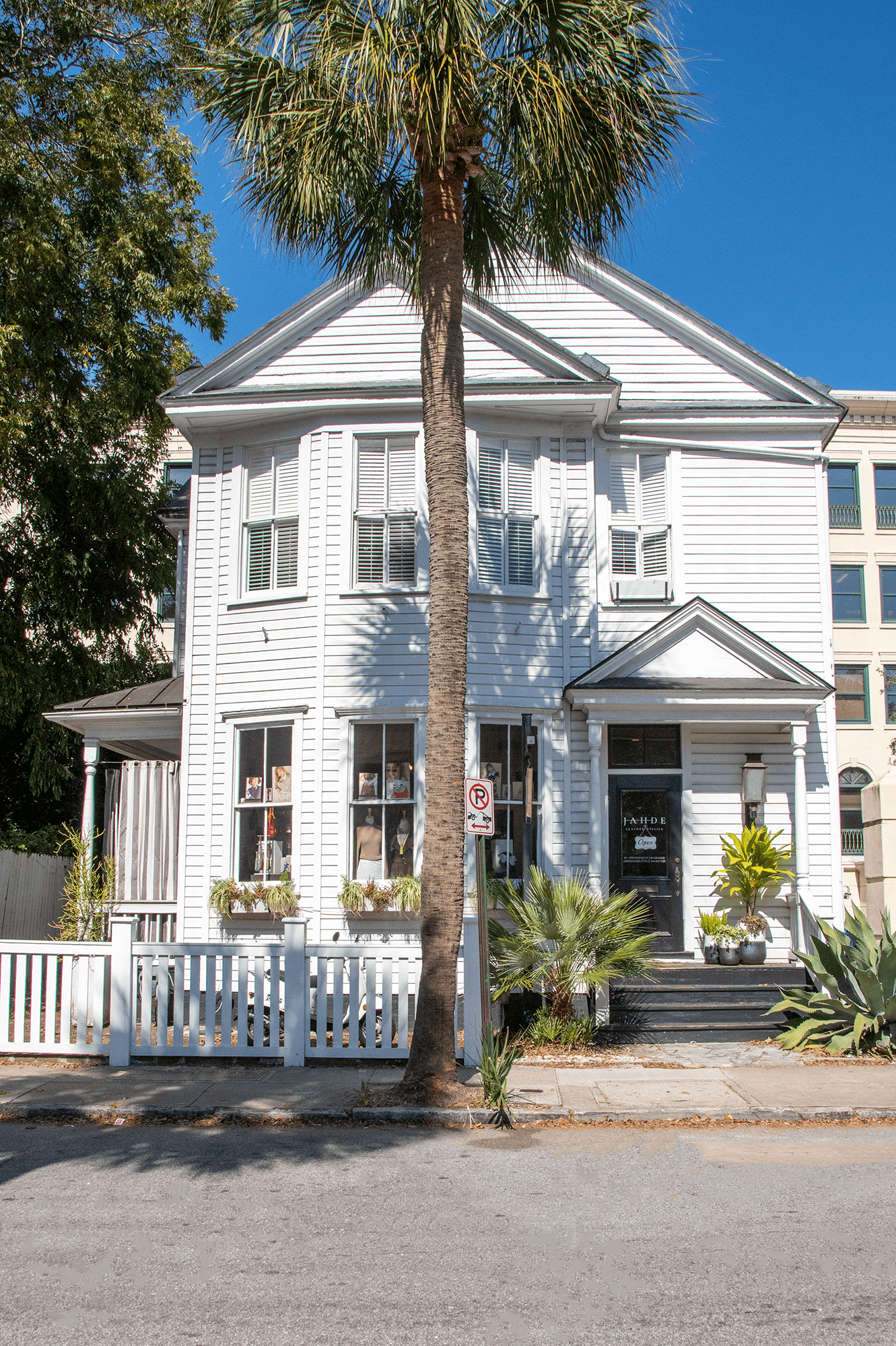 White three-story house with a porch, surrounded by plants and a white picket fence, under a clear blue sky.