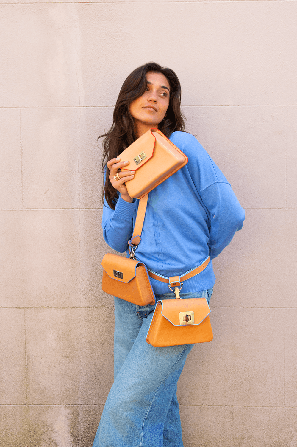 Young woman with long dark hair wearing a blue jacket and jeans, holding three orange bags, standing against a plain wall.