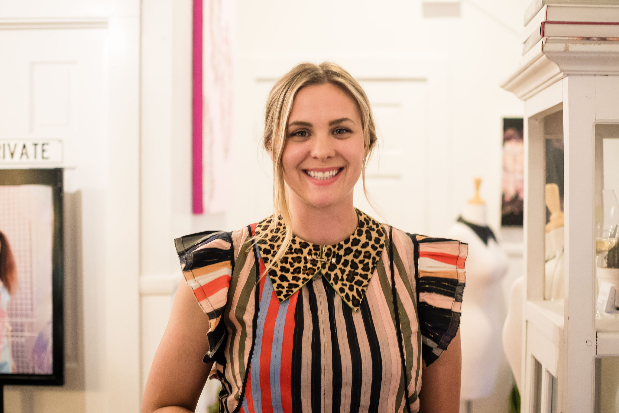 Young woman with blonde hair smiling, wearing a striped top with a leopard print collar, in a bright room.