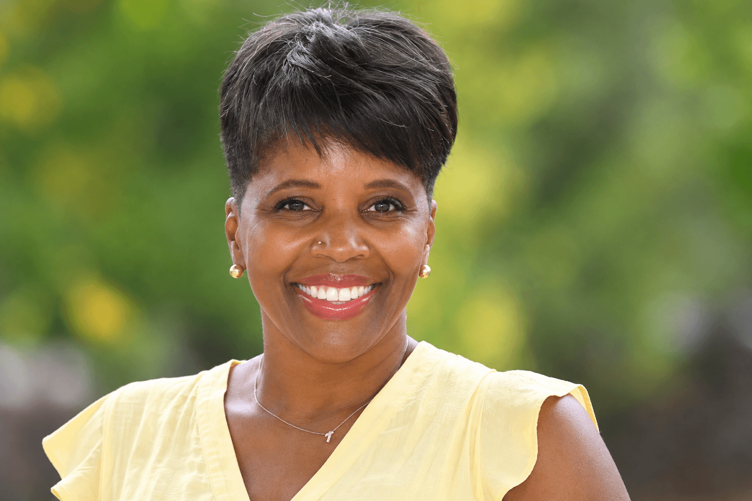 Smiling woman with short black hair wearing a yellow top outdoors, blurred green background.