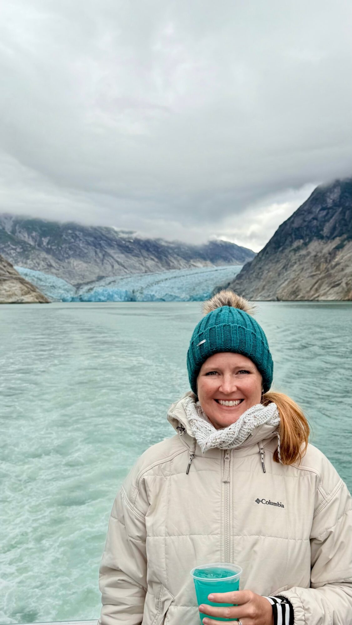 A person in a teal knit hat and beige jacket stands beside a glacial lake, with mountains behind them.