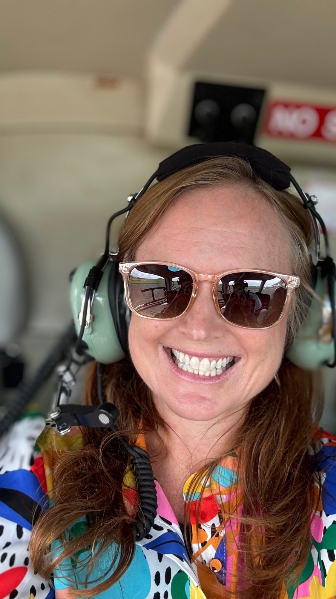 Smiling person wearing sunglasses and a headset inside an aircraft cabin.