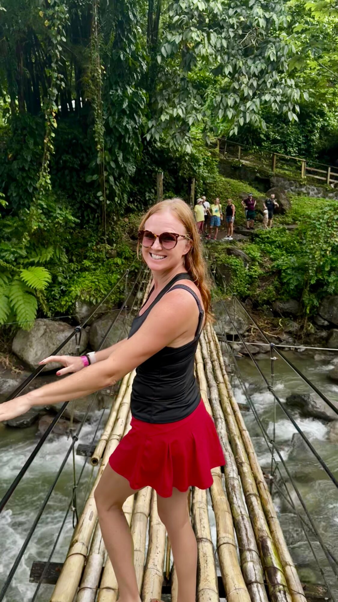Person wearing sunglasses stands on a bamboo footbridge over a stream, with dense greenery and people in the background.