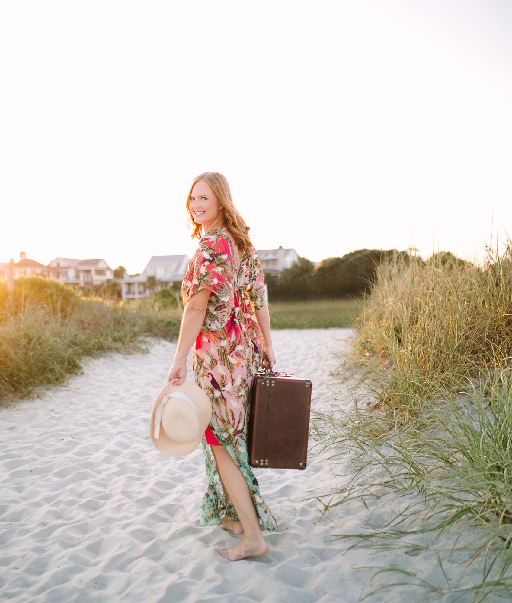 A person in a floral dress walks along a sandy path carrying a hat and suitcase, with beach grass and houses behind her.