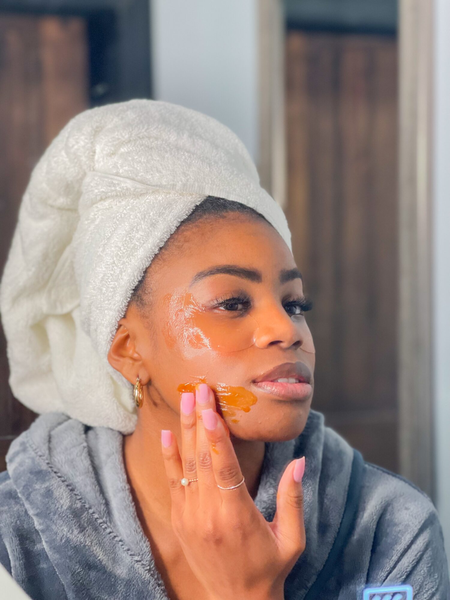 Person wearing a towel turban and gray robe, applying an orange skincare product to their cheek.