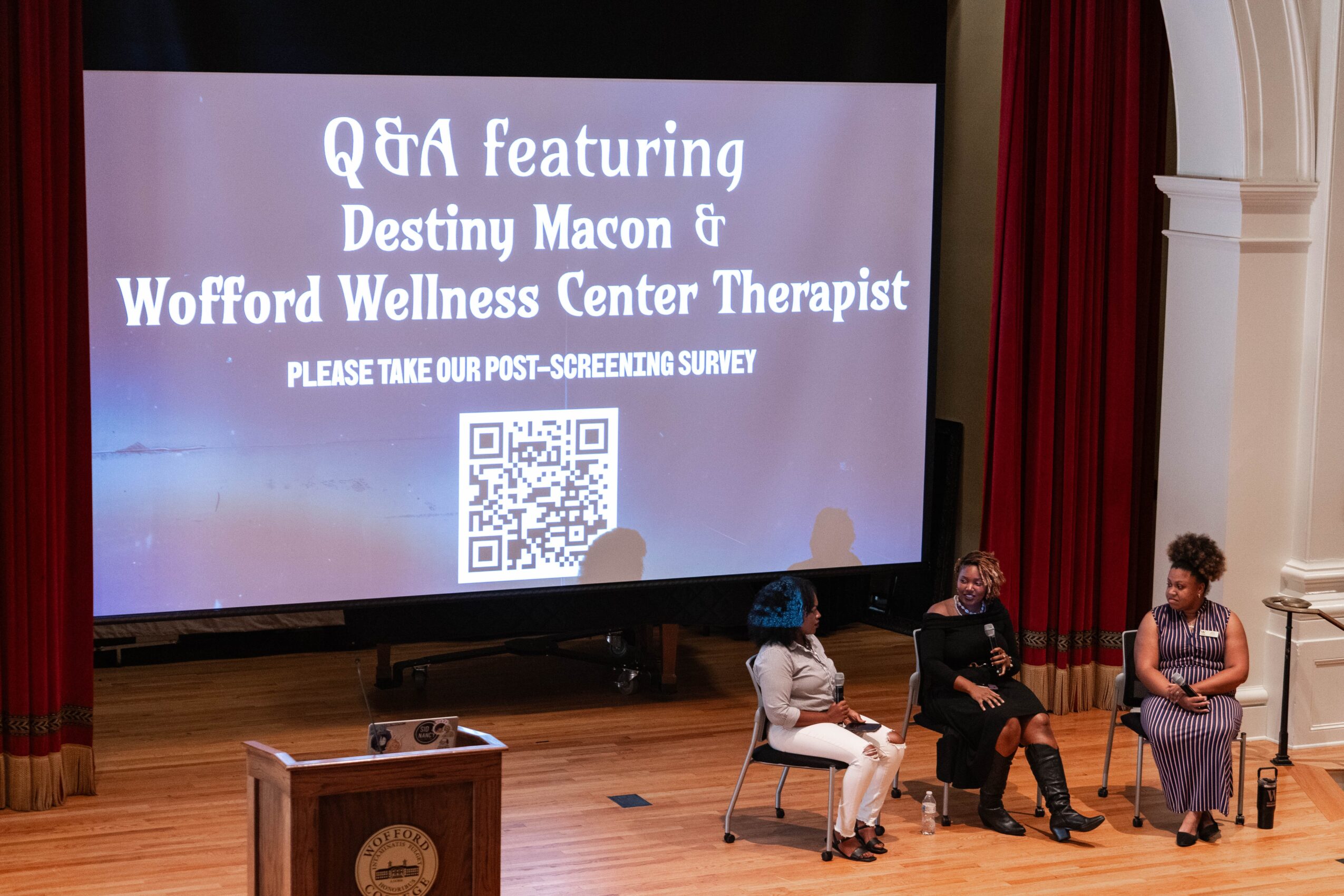 A projected Q&A slide stands on a stage with three seated people, red curtains, and a wooden lectern.