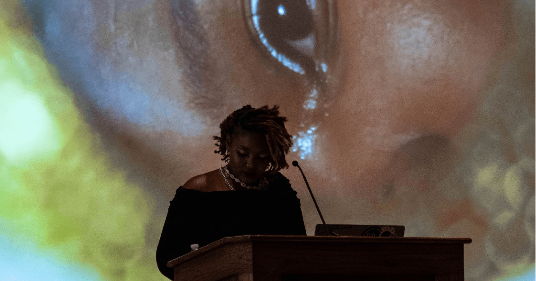 A person speaks at a podium beneath a large projected close-up of an eye.