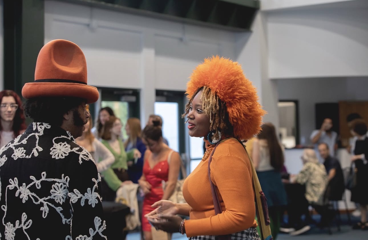 Two people converse at an indoor event; one wears an orange hat, and the other has bright orange hair.