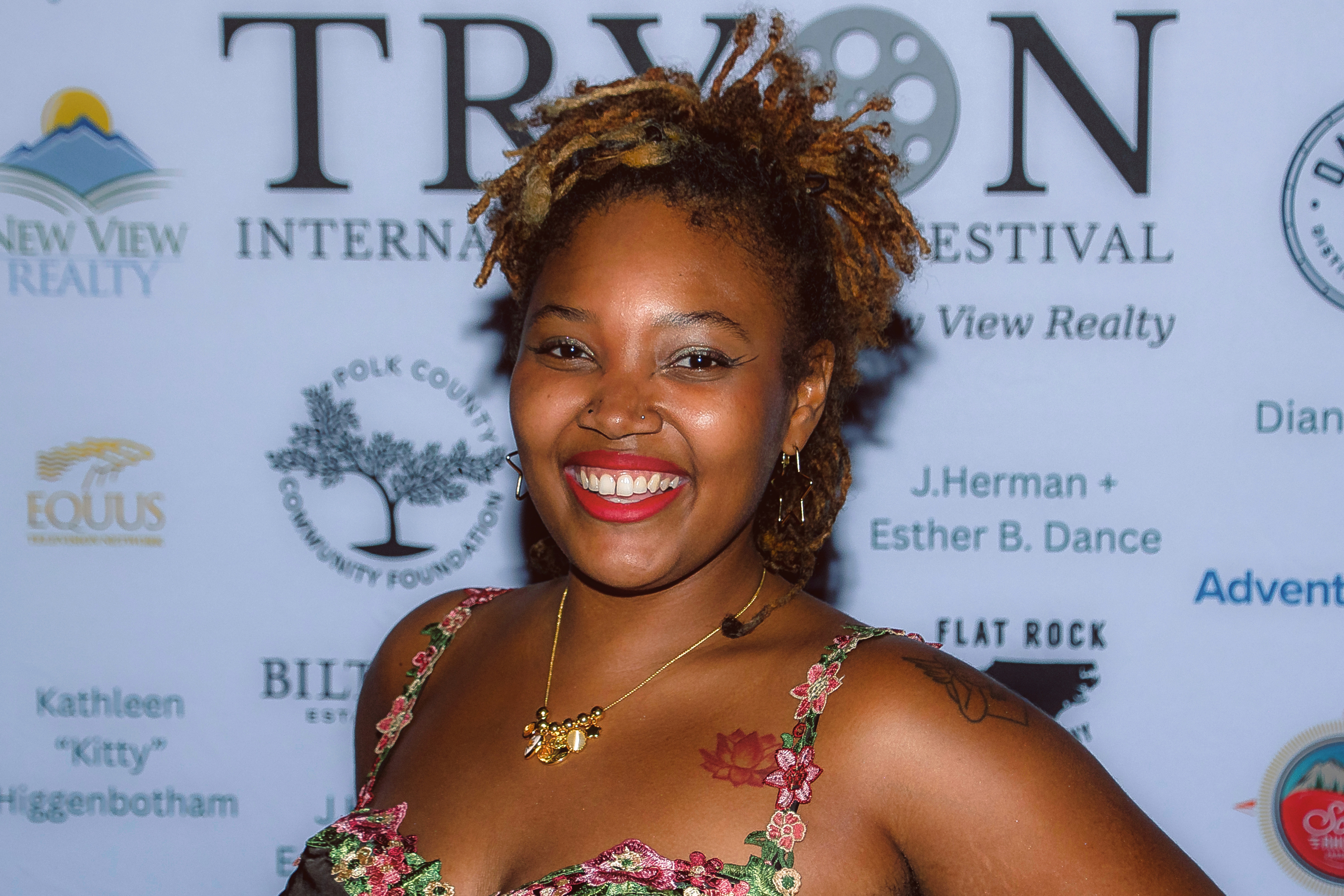 A smiling person with curly hair wearing a floral dress poses in front of an event sponsor backdrop.