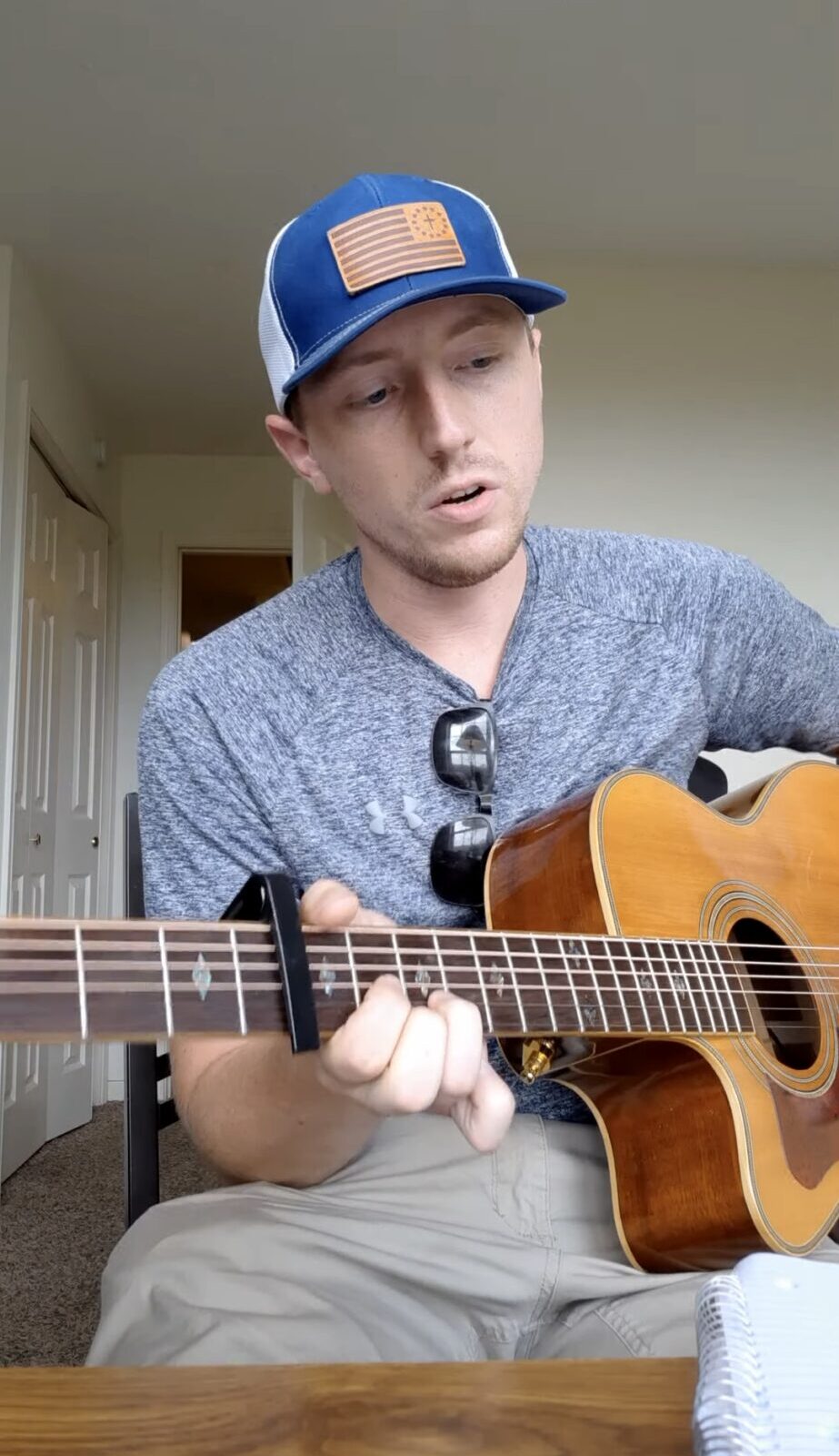 A seated person wearing a blue cap plays an acoustic guitar indoors.