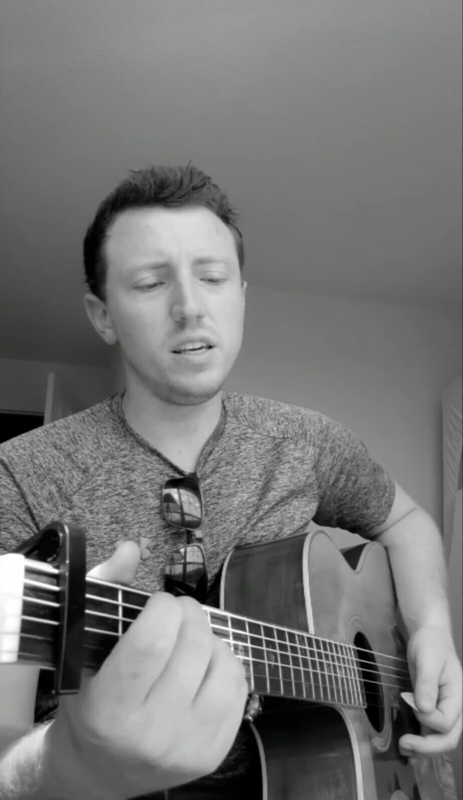 Person playing an acoustic guitar indoors, with a pair of glasses hanging from their shirt.