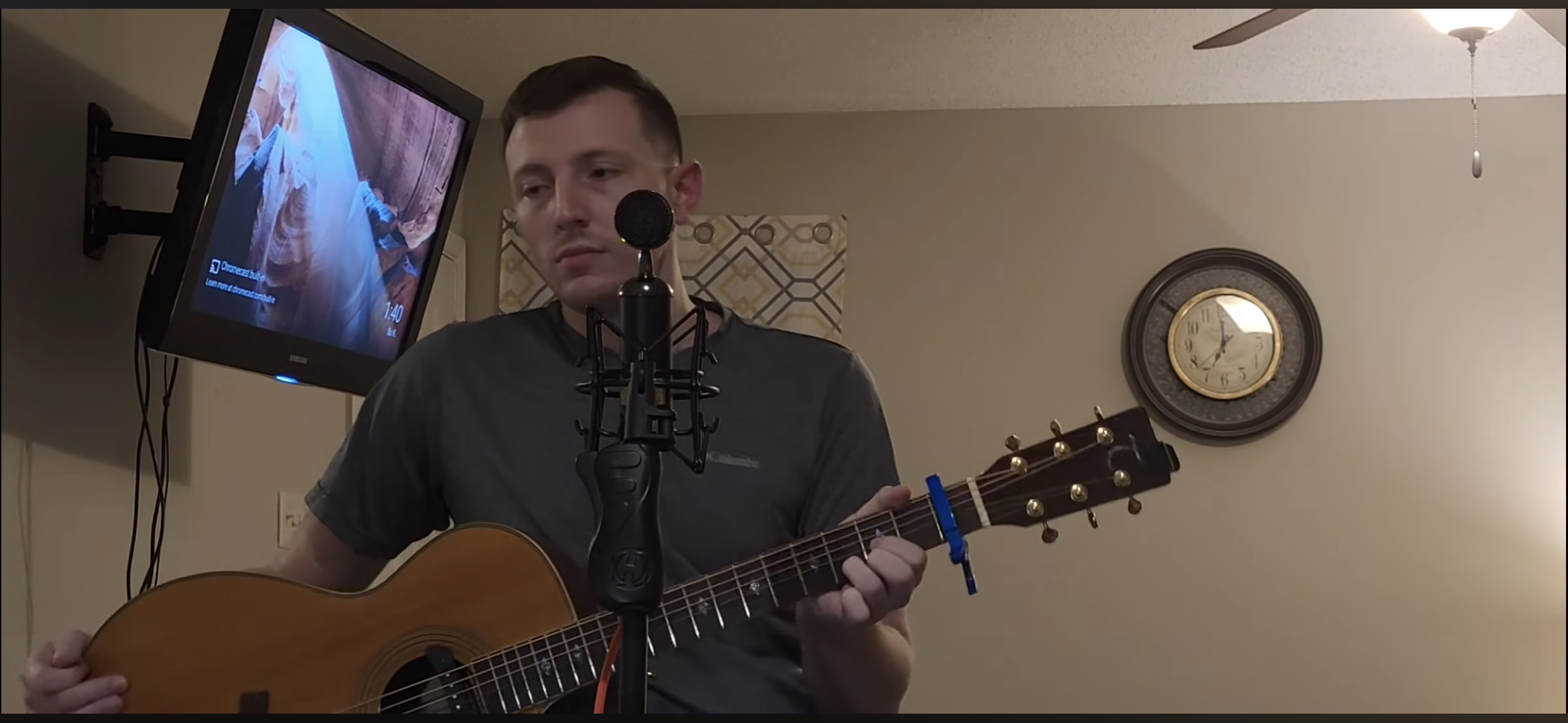 A person holds an acoustic guitar beside a microphone, with a television and wall clock in the background.