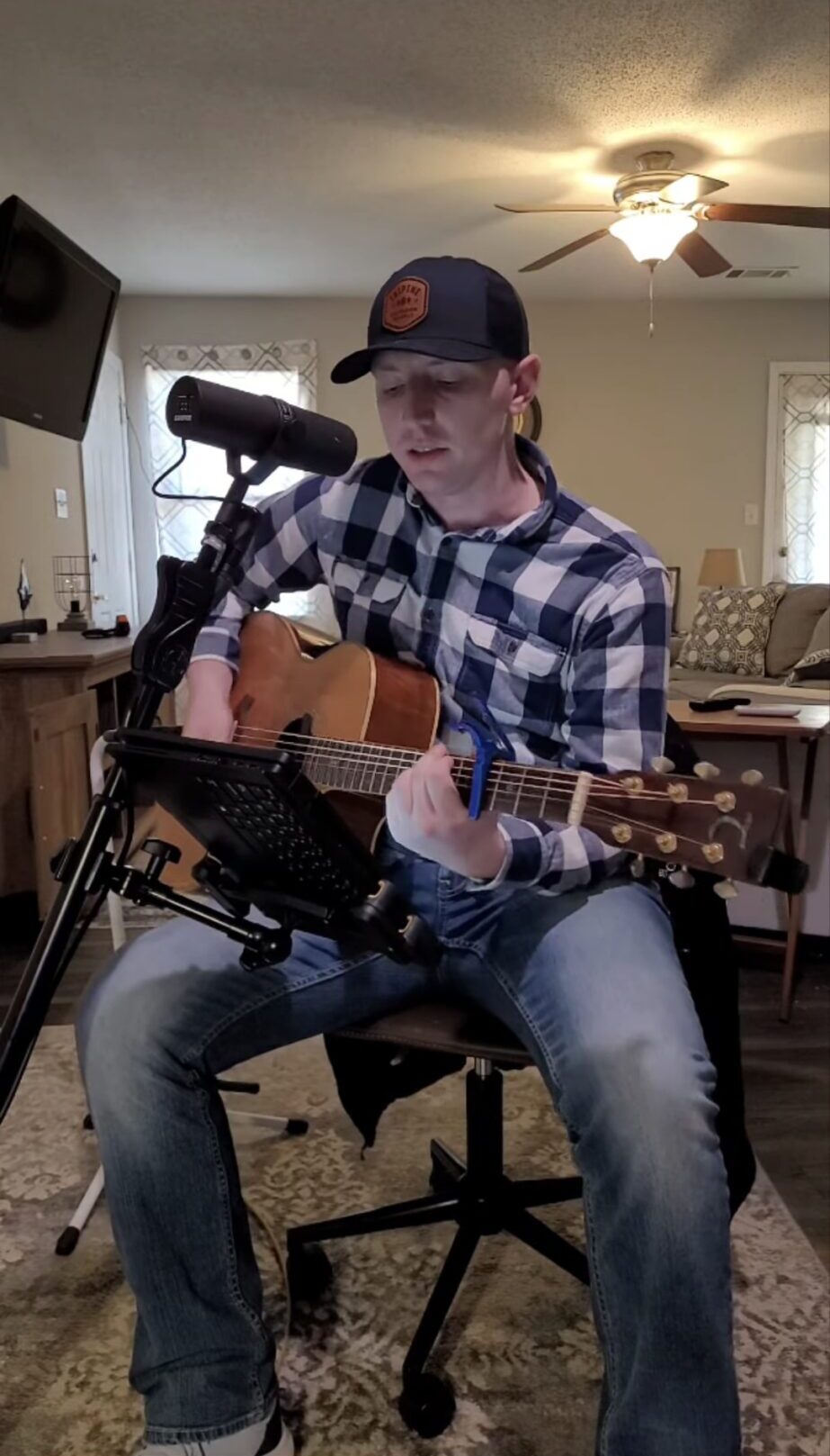 A seated person plays an acoustic guitar near a microphone in a living room. Subtitles appear along the bottom.