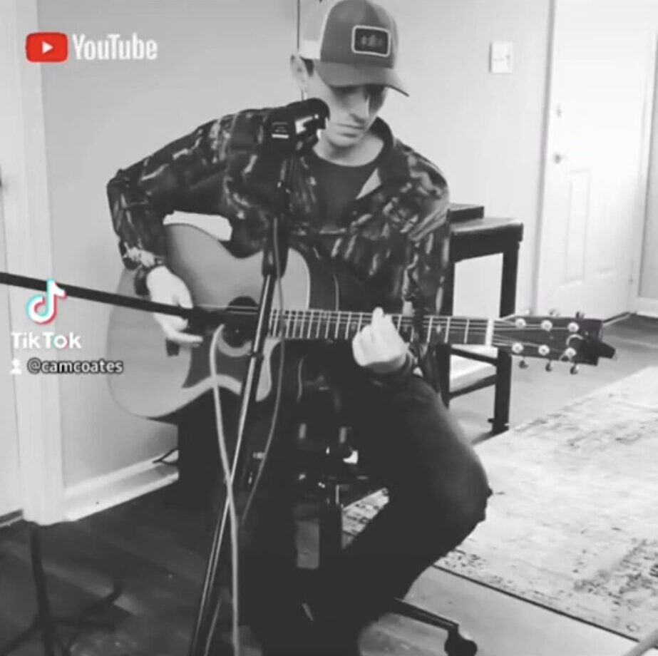 A seated person wearing a cap plays an acoustic guitar indoors.