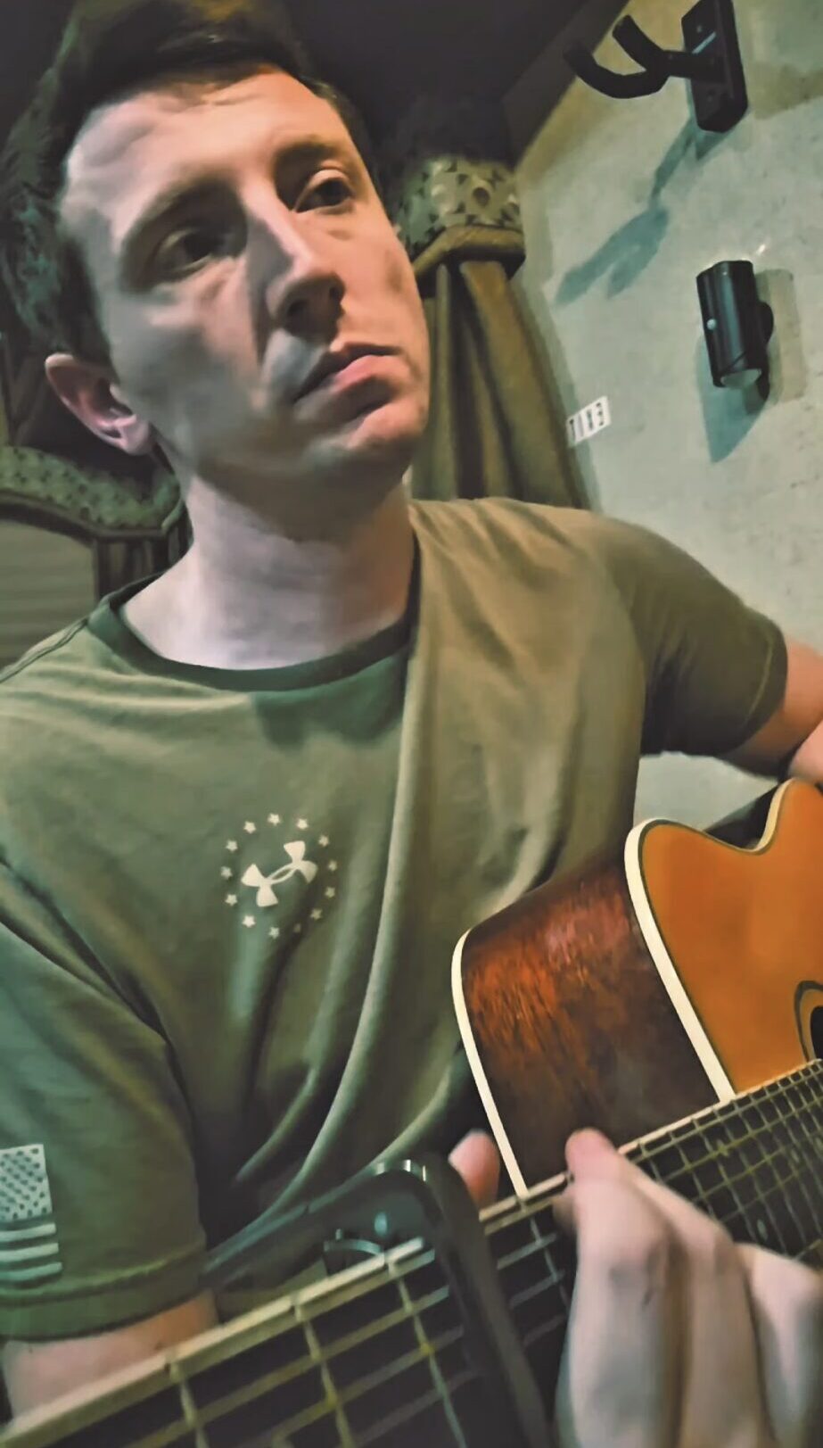 A person in a green shirt holds and plays an acoustic guitar indoors.