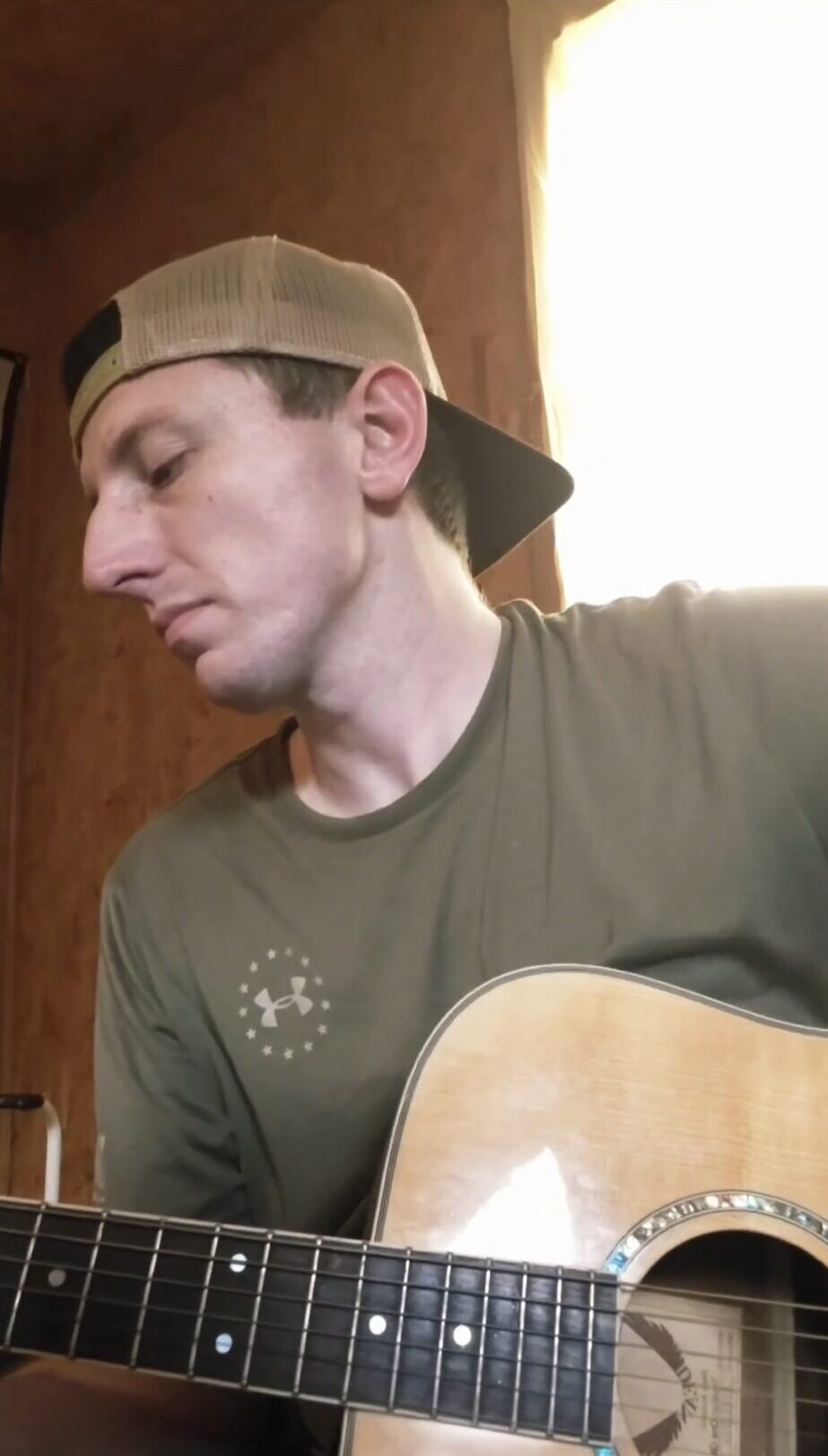 A person wearing a backward cap holds an acoustic guitar indoors beside a bright window.