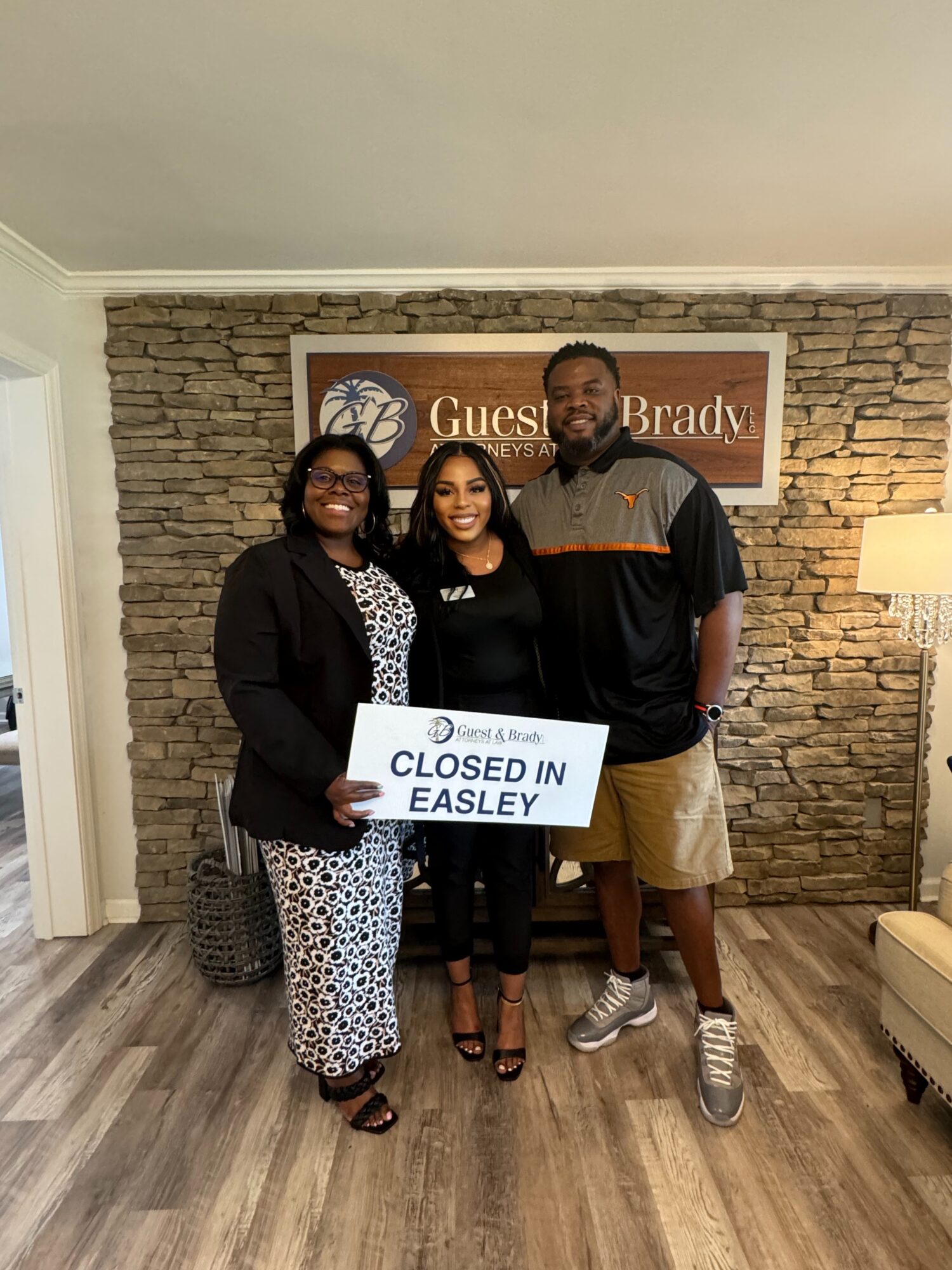 Three people stand in front of a stone wall, holding a sign that reads “Closed in Easley.”