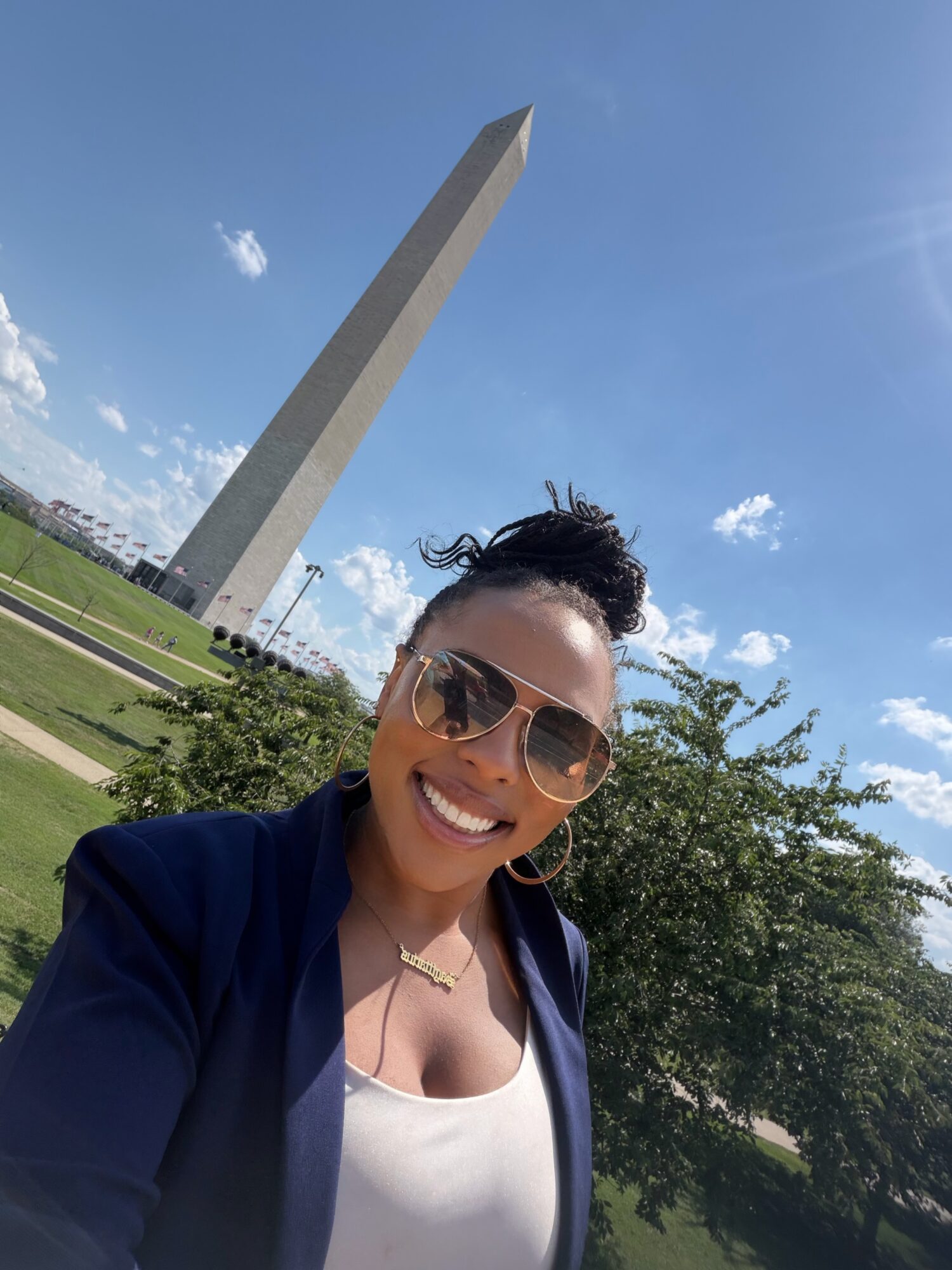 Person wearing sunglasses and a navy jacket smiles in front of a tall stone monument and trees.