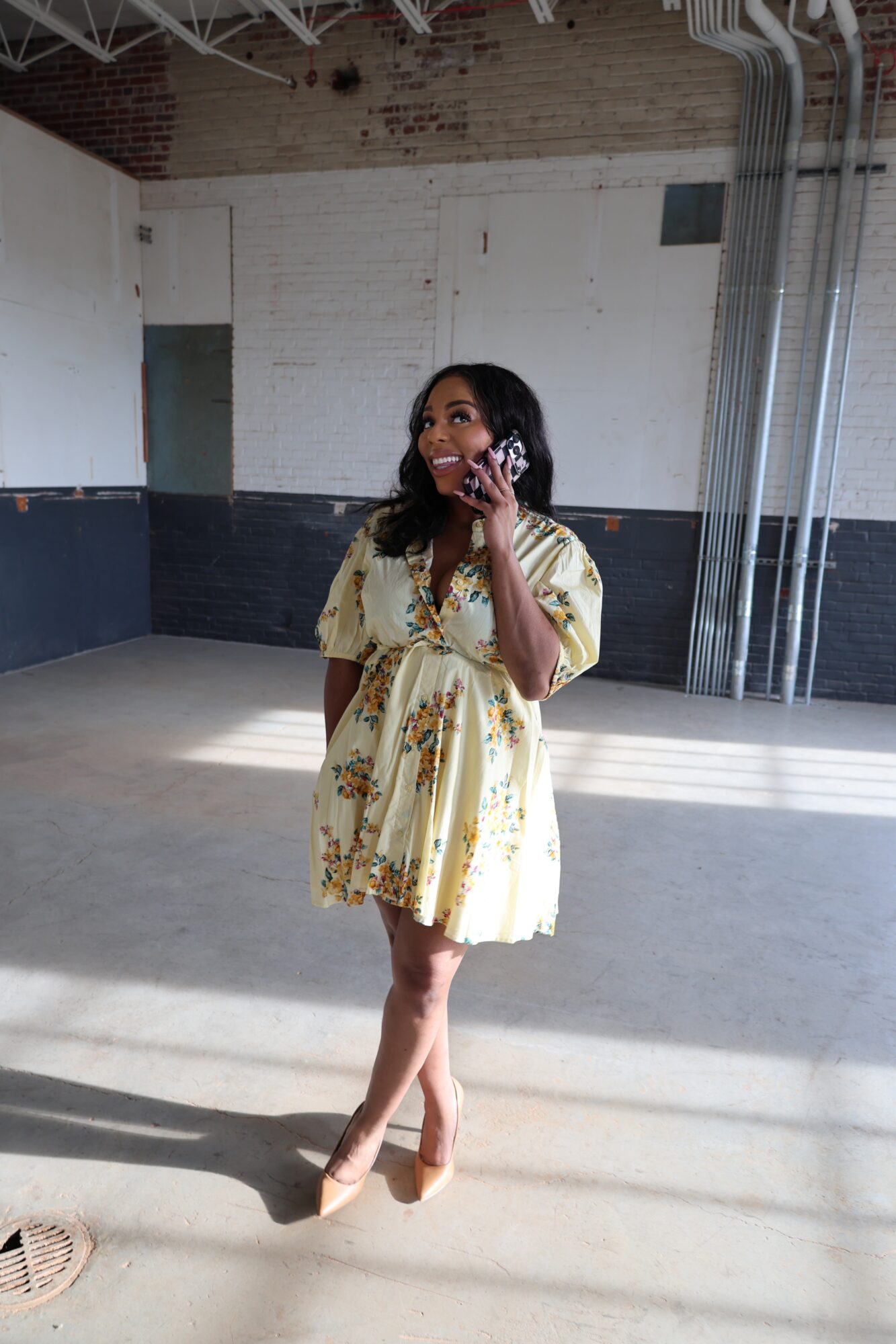 A person in a floral dress stands in a large unfinished room, holding a phone to their ear.