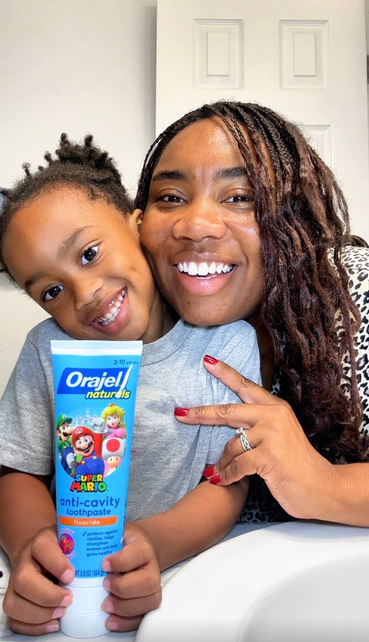 A smiling adult and child pose together, holding a tube of Orajel near a white door.