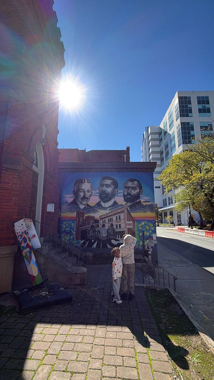 Two people stand before a large mural on a brick wall beside city buildings on a sunny day.
