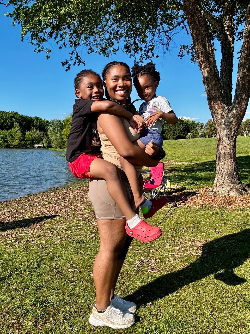 A person carries two children beside a lake under a tree.