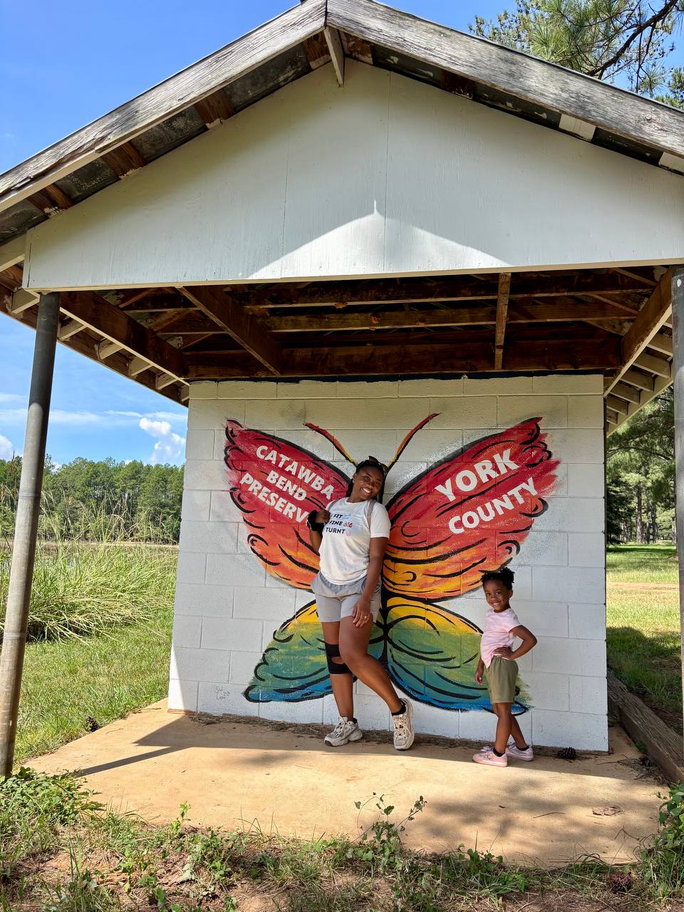 Two people pose in front of a painted butterfly-wing mural beneath a wooden pavilion.