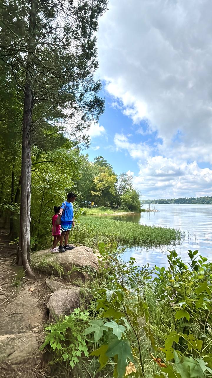 Two people stand beside a lakeshore path, surrounded by trees and shoreline plants beneath a cloudy sky.