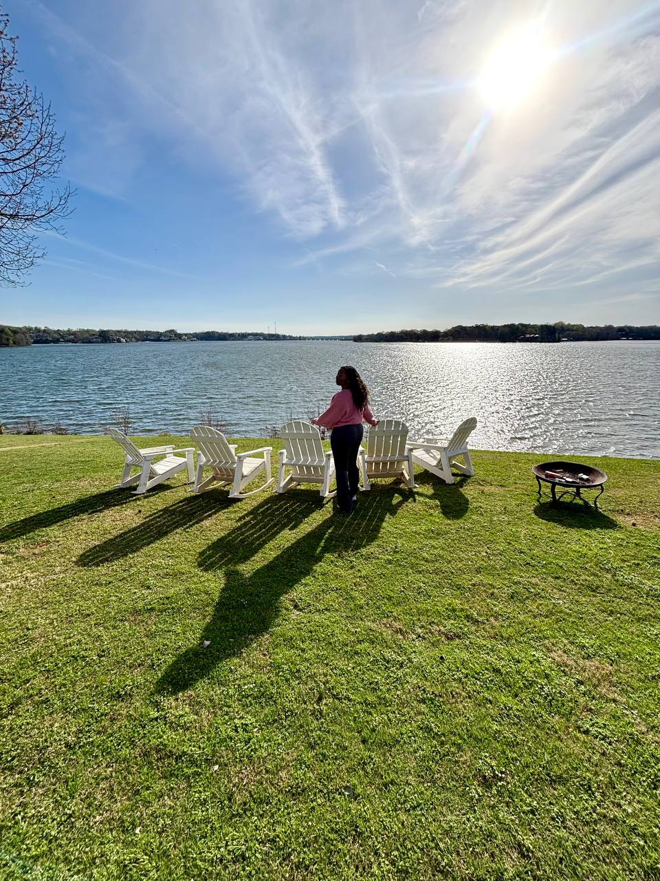Person standing beside white chairs on a grassy lakeshore, with a fire pit and bright sun overhead.