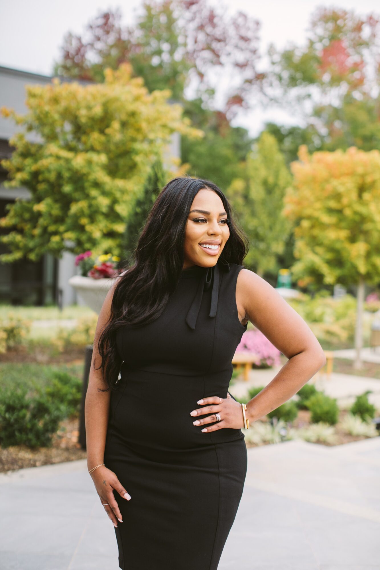 A person in a black sleeveless dress poses outdoors beside landscaped gardens and autumn-colored trees.