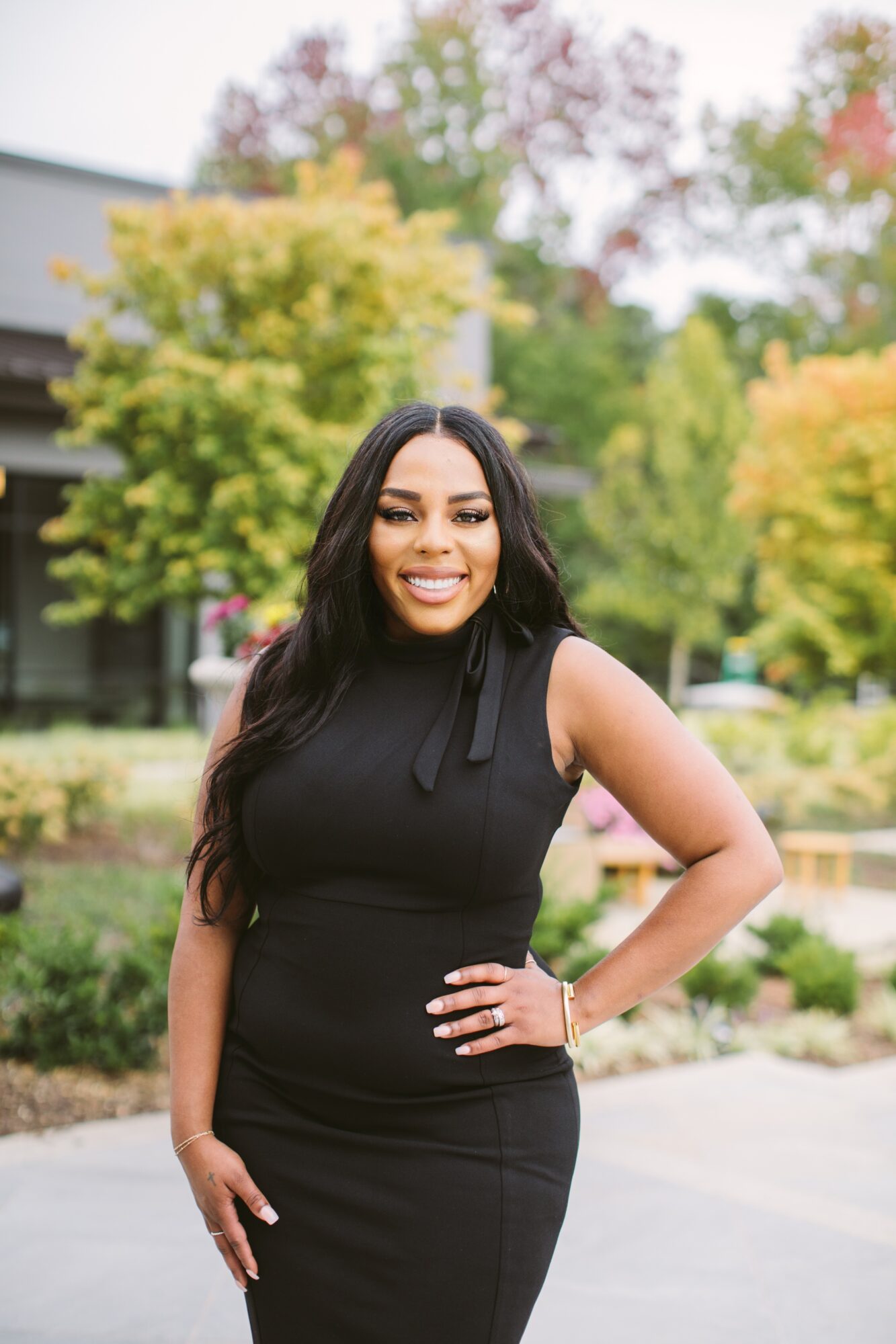 Person in a black dress standing outdoors with one hand on her hip, trees and a building behind her.